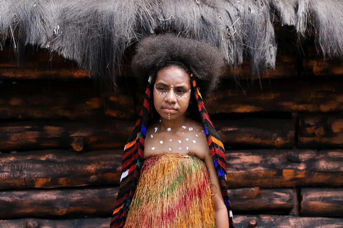Woman in vibrant traditional attire with facial paint against rustic background.